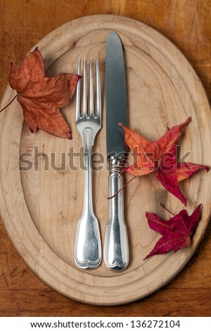 Silver fork and knife on a rustic wood plate with colorful autumn leaves. Top view, looking down.