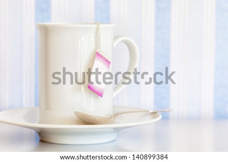 White cup and saucer with a silver spoon and a blank tea bag tag hanging over the side. Blue and white striped background.