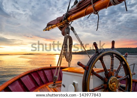 Sunrise at sea on a tall ship classic sailboat.  Close up of the wheel, boom and stern against a dramatic sky and gold sunlight reflections. Concepts: Serenity, prosperity, optimism, positive, future