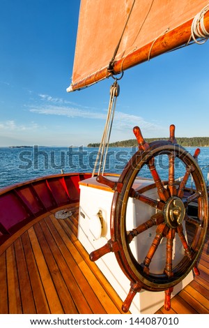 Smooth sailing on a classic schooner with a close view of the captain\'s wheel. Colorful image with a bright blue sky, an orange sail and wood decking.