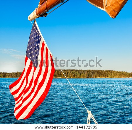 American flag flying from a sailboat off the coast of Maine, USA. The flag and sail frame a view across the water of a forested island in Penobscot Bay. Bright blue sky with copy space.