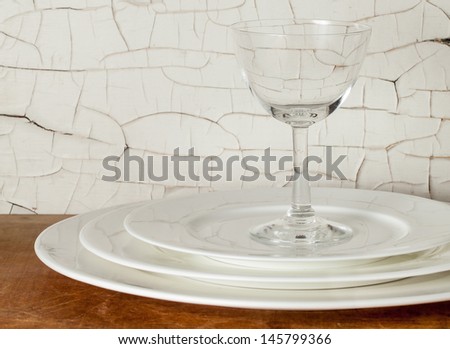 White porcelain dinner plates stacked with a wine glass against a distressed wall with cracked white paint. Fine china in a grunge setting.