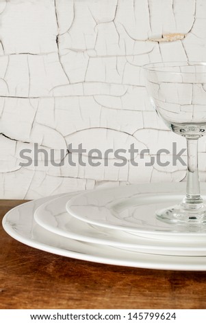White porcelain dinner plates stacked with a wine glass against a distressed wall with cracked white paint. Fine china in a grunge setting. Vertical with copy space.