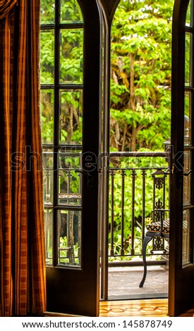 French doors open onto a balcony with a view of leafy green trees. Elegant silk drapery in tones of orange and rust.
