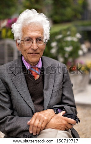 Portrait of a happy senior man with white hair wearing a pink shirt with a tie and jacket, sitting outside in a garden. Shot with shallow dof.