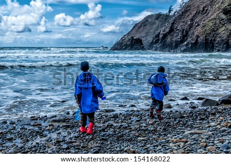 Two children wearing rain jackets and rubber boots play on a rocky beach in stormy weather. Dramatic clouds overhead.