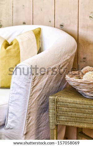 Chair in a white slipcover with a striped pillow next to a green wicker side table with a basket of shells. Comfortable, casual seaside cottage decor.