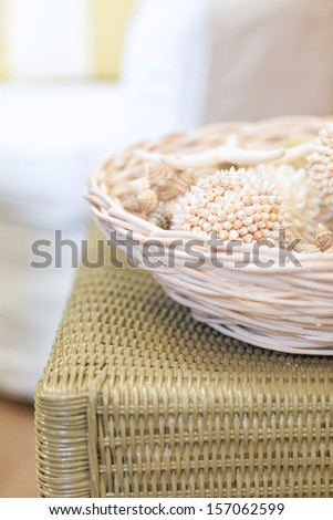 Basket of decorative shell balls on a painted wicker side table. Shabby chic seaside cottage decor.  Selective focus with creamy background blur.