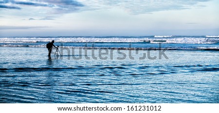 Photographer at work with a tripod and camera stands in the ocean waves to catch a picture. Concept for determination and perseverance.