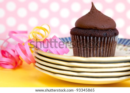 Chocolate cupcake with chocolate frosting on a pink and white polka dot background with a stack of vintage plates