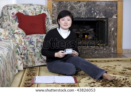 A pretty Asian woman sitting on carpet floor nearby fireplace and drinking tea