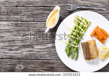 grilled hake served with asparagus, piece of lemon, baby carrots, and the hollandaise sauce on the white platter on the old rustic table, view from above