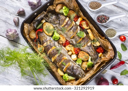 delicious trout fishes baked with potatoes, broccoli, lemon, tomatoes and spices in baking dish on a wooden background, view from above