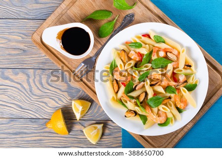 tasty penne pasta salad with shrimps, mussels and baby spinach leaves on a white dish with vintage fork, caramelized balsamic vinegar in a sauce boat and lemon slices on an rustic table, top view