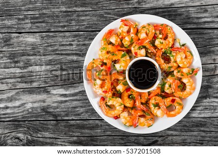 butter garlic fried shrimps sprinkled with pieces of chilli and parsley on white dish with soy sauce in small bowl in centre, on wooden table,view from above