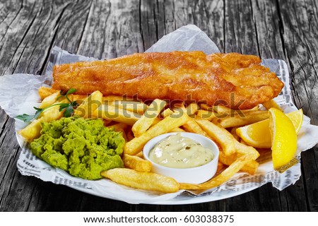 delicious crispy fish and chips - fried cod, french fries, lemon slices, tartar sauce and mushy peas on plate on paper, on wooden table, front view from above, close-up