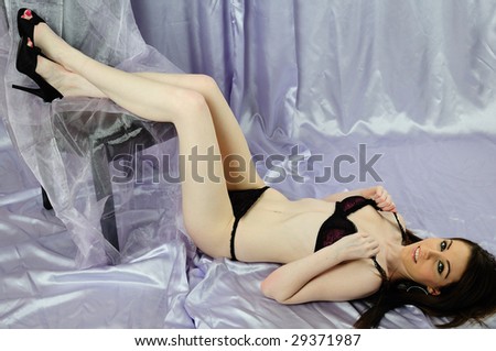 female lingerie model lying on the floor on  a lilac back drop floor length. Wearing black lingerie with her feet up on a leather high back chair.