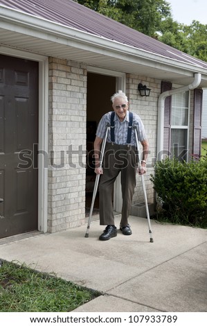 An elderly disabled veteran with crutches, with a smiling happy face, standing alone outside of his tiny apartment.