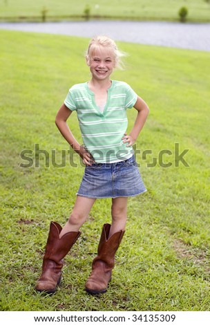 A cute little young cowgirl with blonde hair, wearing boots on a farm with shallow depth of field