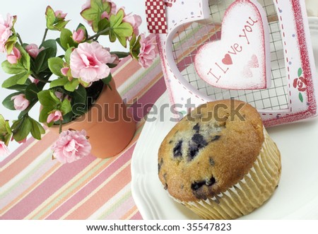A delicious blueberry muffin on a plate with a pink feminine setup and a heart with I Love You written on it, diagonal viewpoint