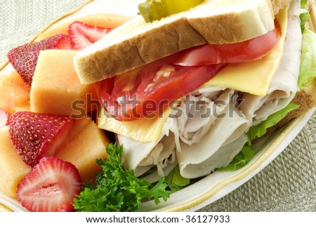 A delicious turkey sandwich filled with sliced turkey, tomatoes, cheese and lettace on toasted bread with fresh strawberries and cantaloupe, diagonal viewpoint, closeup