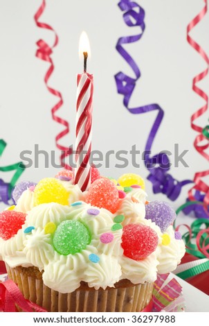 A colorful birthday cupcake, decorated with gumdrops and sprinkles, with one lit birthday candle, vertical with shallow depth of field