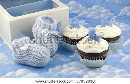 A pair of crocheted blue baby booties, a gift box and delicious chocolate cupcakes with vanilla frosted and baby carriage decoration, baby shower gifts, vertical with copy space