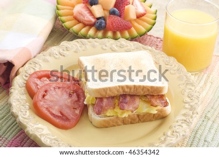 A scrambled egg and bacon sandwich with sliced tomatoes, fresh fruit and orange juice, served on a yellow plate with napkin and placemat, horizontal