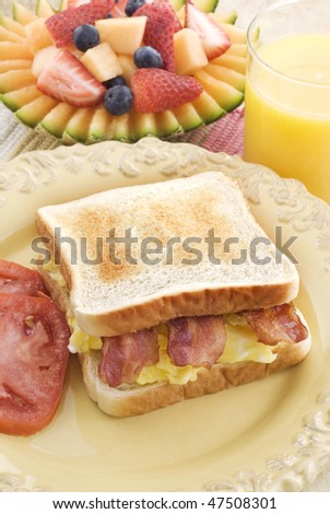 A bacon and scrambled egg sandwich on toasted bread with slice tomatoes, fresh fruit, and orange juice, vertical with copy space