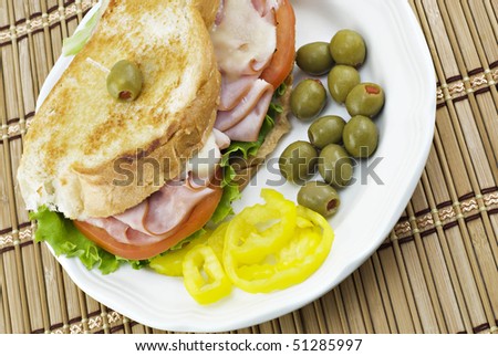 A grilled ham and swiss cheese sandwich on a plate with olives and banana peppers on a bamboo placemat, horizontal with copy space