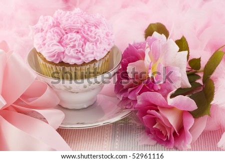 A pretty pink cupcake in a teacup surrounded by pink boa, bow, and pink roses, horizontal with copy space