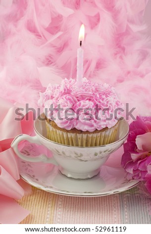 A pink birthday cupcake with one lit candle in a teacup with pink background, vertical, low key image, copy space