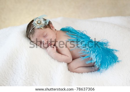A studio portrait of a beautiful one week old baby girl sleeping, with a blue feather and flower headpiece