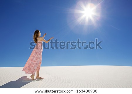 young woman in long dress standing in white sand desert with praising hands towards the sun