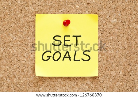 Set Goals on yellow sticky note pinned with red push pin on cork board.