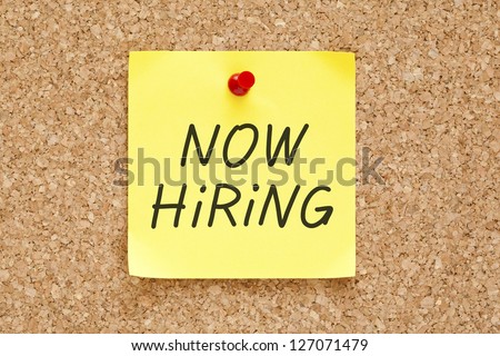 Now Hiring written on an yellow sticky note pinned with red push pin on cork bulletin board.