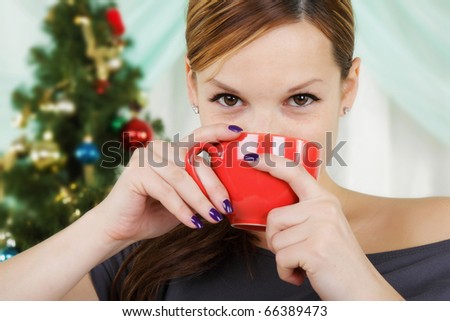 The young beautiful woman sits near a christmas fur-tree with a red cup