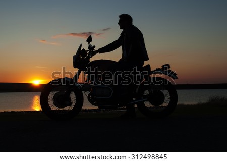 Motorbiker in silhouette at sunset at lake man on motorbike one hand on handlebar