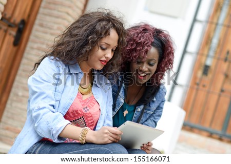 Portrait of two beautiful girls with tablet computer in urban background, black and mixed women. Friends talking