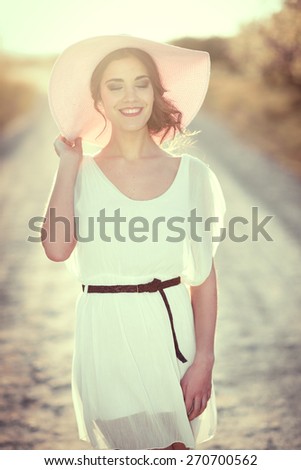 Portrait of young woman with eyes closed in a rural road in the spring time. Girl wearing white dress, brown belt and pink sun hat. Backlight photography with the Sun in the back of the woman.