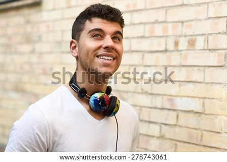 Portrait of young man in urban background smiling with headphones. Wearing white t-shirt near a brick wall