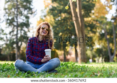 Young happy Woman in checkered shirt and blue jeans with toothy Smile and sunglasses. Blonde girl drinking coffee in park sitting on grass wearing casual clothes smiling