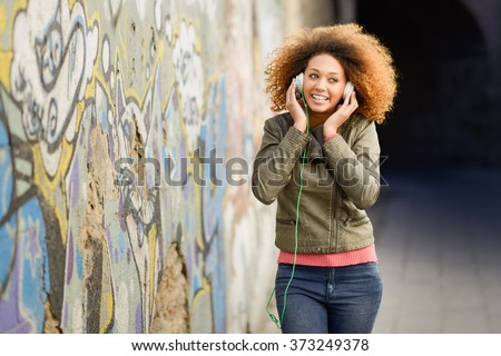 Portrait of young attractive black girl in urban background listening to the music with headphones. Woman wearing leather jacket and blue jeans with afro hairstyle