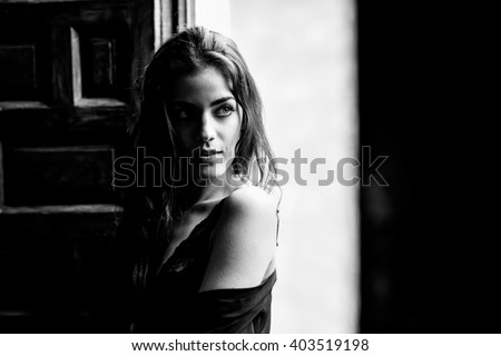 Sexy young woman in lingerie standing near a window in her bedroom. Brunette girl wearing black body and neglige. Female in underwear. Black and white photograph.