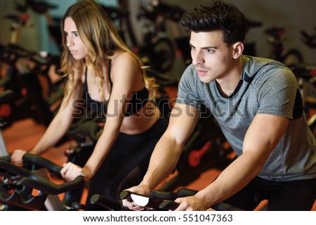 Young man and woman biking in the gym, exercising legs doing cardio workout cycling bikes. Two people in a spinning class wearing sportswear.