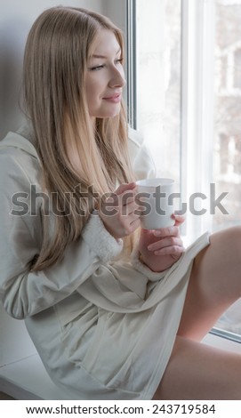 A happy young girl in a short dressing gown is sitting with mug near the window