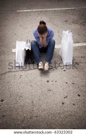 A casually dressed young woman sitting in an empty parking lot with shopping bags and looking down as she covers her face.  Maybe she is feeling down because she spent more than she intended to.