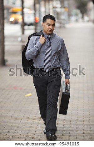 Stock photo of a Hispanic businessman walking down the sidewalk in an urban business district carrying a coat over one shoulder and his breifcase.