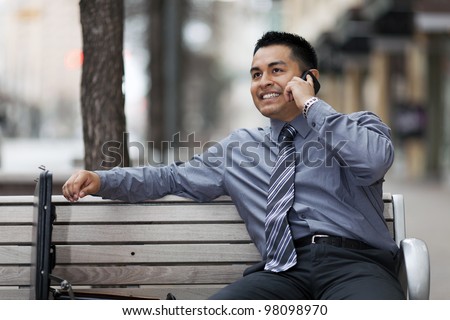 Stock photo of a smiling Hispanic businessman sitting on a city bench in a business district with an open briefcase, while he talks on a cell phone.
