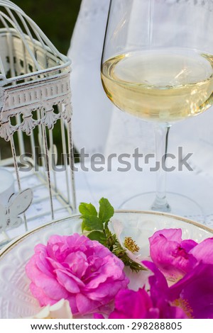A summer still life. A glass of white wine, a white cage and pink rose hip flowers on the white porcelain plate. Outdoor.